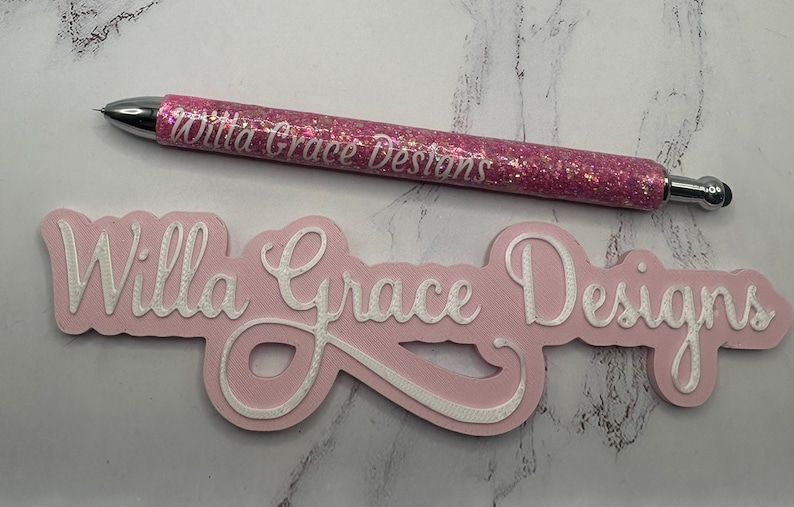 May include: Pink glitter pen with the text "Willa Grace Designs" on it, next to a pink sign with the same text in white raised lettering.
