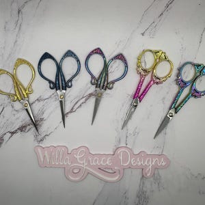 May include: Five pairs of decorative scissors with intricate designs. The scissors are in various colors, including gold, blue, and purple. The scissors are arranged in a row on a white marble surface. The text "Willa Grace Designs" is visible in the lower center of the image.