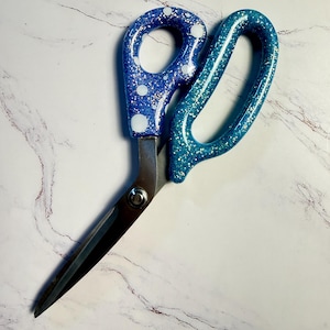 May include: A pair of scissors featuring metallic silver blades and handles coated in a glittery finish. The smaller handle is dark blue with white polka dots, while the larger handle is a bright turquoise blue, both embedded with fine sparkles.