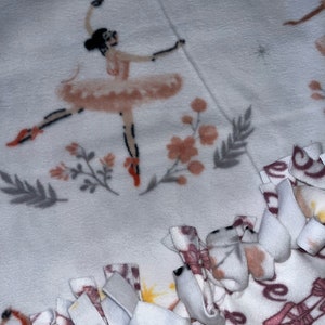 May include: White fleece blanket featuring a ballerina in a pink tutu, pointe shoes, and a black bun. The blanket has a tied edge and a pattern of ballet shoes and floral accents in pink and gray.