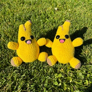 May include: Two crocheted yellow banana-shaped plush toys with smiling faces and black eyes. The toys are sitting on a bed of green grass.