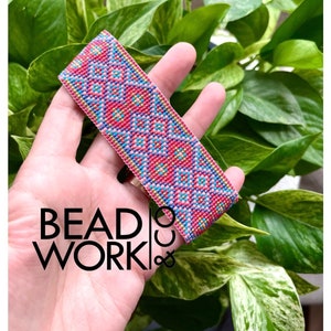 May include: A pink, blue and gold beaded bracelet with a geometric pattern. The bracelet is held in a hand against a green leafy background.