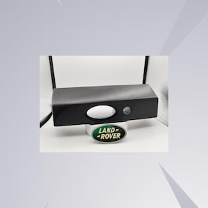 Pode incluir: Uma peça automotiva preta com uma abertura oval e um pequeno botão circular. A peça é exibida em uma superfície branca com o logotipo da Land Rover em verde e branco.