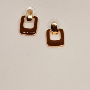 May include: A pair of gold-toned earrings with a square shape and a smaller square cutout in the centre. The earrings have a shiny finish.