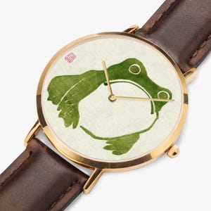 May include: A wristwatch with a gold-tone circular case and a dark brown leather strap. The watch face features a cream-coloured background with a green watercolour-style illustration of a frog and thin gold-tone hour, minute, and second hands.