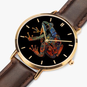May include: A gold-coloured wristwatch with a brown leather strap. The black watch face features a colourful frog illustration in shades of orange, blue, and green. The watch has gold hands and hour markers.