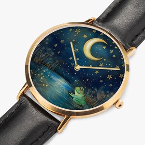 May include: A gold-coloured wristwatch with a black leather strap. The watch face depicts a frog in a pond beneath a crescent moon and starry night sky. The watch hands are gold.