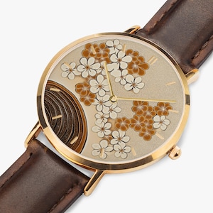Op de afbeelding: Een gouden polshorloge met een bruin leren bandje. De wijzerplaat van het horloge heeft een bloemenmotief met witte en bruine bloemen.