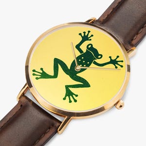 Puede incluir: Un reloj de pulsera con caja redonda en tono dorado y correa de cuero marrón oscuro. La esfera presenta un fondo amarillo brillante con un gráfico de una rana en verde oscuro en el centro. Tres finas manecillas doradas marcan la hora.