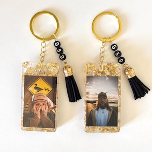 May include: Two gold keychains with black tassel charms. Each keychain features a rectangular resin charm with gold leaf accents and a photo inside. One charm has a photo of a person wearing a hat and the other charm has a photo of a person wearing a hat and sunglasses. The keychains also have black beads with white letters that spell "G 5 9".