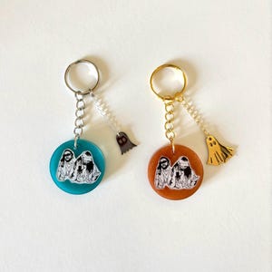 May include: Two keyrings with circular pendants showing a black and white image of two people. One has a teal pendant and silver hardware, the other has a copper pendant and gold hardware. Each has a ghost charm.