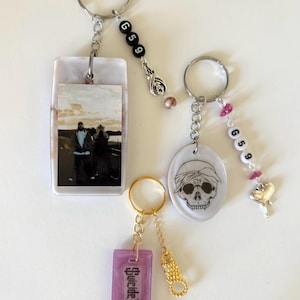 May include: Three keychains with different charms. The first keychain has a white and pink marbled rectangle with a photo of two people. The second keychain has a white and pink marbled oval with a black and white skull design. The third keychain has a purple rectangle with the text "Suicide Boy$".