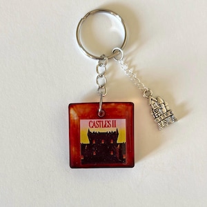 Peut inclure: Un porte-clés avec un pendentif carré brun-rouge représentant un château et le texte "CASTLES II". Un petit charm en argent en forme de château est suspendu à la chaîne. Le porte-clés a un anneau argenté.