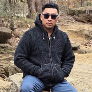 May include: A man wearing a dark gray hooded sweater with a zipper and a pair of blue jeans. He is wearing blue-framed sunglasses.