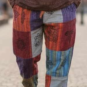 May include: Multi-coloured patchwork trousers in red, orange, blue, and grey with embroidered patterns. The trousers have a drawstring waist and are made of a soft, textured fabric. The person is walking barefoot on a brick surface.
