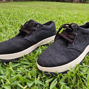 Pode incluir: Um par de t&ecirc;nis baixos de lona pretos com solas brancas. Os sapatos t&ecirc;m cadar&ccedil;os pretos e est&atilde;o sobre um leito de grama verde.