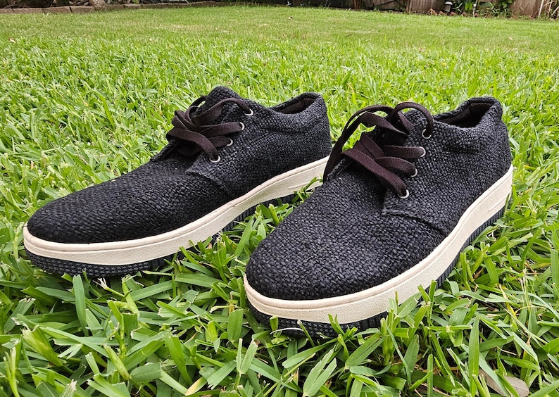Pode incluir: Um par de t&ecirc;nis baixos de lona pretos com solas brancas. Os sapatos t&ecirc;m cadar&ccedil;os pretos e est&atilde;o sobre um leito de grama verde.