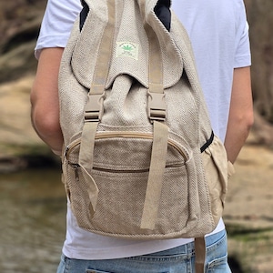 Könnte beinhalten: Ein beigefarbener Hanfrucksack mit einem braunen Reißverschluss und verstellbaren Trägern. Der Rucksack hat ein kleines grünes Blattlogo mit dem Text "Hemptique" und "Home Grown Textiles" darauf.