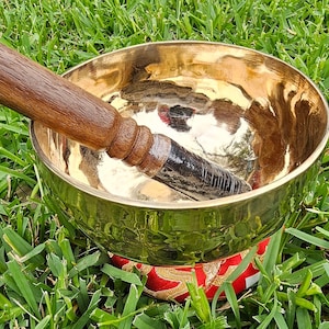 May include: A golden singing bowl with a wooden mallet resting inside. The bowl sits on a red and gold patterned cushion, set against a backdrop of green grass. The mallet is dark brown with a textured grip.