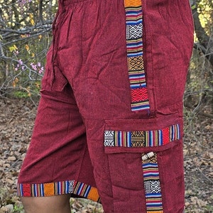 May include: Burgundy cotton shorts with colorful woven trim detail. The shorts have a drawstring waist and a cargo pocket on the right side.