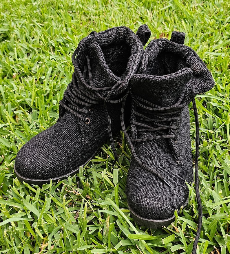 Pode incluir: Um par de botas de cano baixo de lona preta com cadar&ccedil;os. As botas est&atilde;o sobre um leito de grama verde.