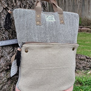 Puede incluir: Una bolsa de mano de cáñamo con estampado de chevron blanco y negro con una sección inferior de estampado de rombos beige. La bolsa tiene un asa de cuero marrón y una cremallera de cuero marrón. La bolsa tiene un pequeño borla negro en el lateral. La bolsa tiene una etiqueta que dice "GANAMO, 100% CÁÑAMO, FIBRA NATURAL".