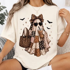 May include: A cream-colored t-shirt featuring a graphic of a ghost wearing a leopard print bow and heart-shaped sunglasses. The ghost is holding a leopard print tote bag and a coffee cup with a smiley face, surrounded by small black bats and stars.
