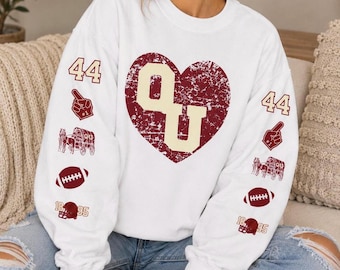 May include: A white long-sleeve crewneck sweatshirt featuring a large distressed maroon heart on the chest with the letters OU in cream. Both sleeves display repeating graphics of the number 44, a foam finger, a covered wagon, a football, and a helmet.