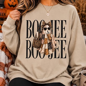 May include: A beige crewneck sweatshirt featuring a graphic of a ghost wearing a leopard print bow, heart-shaped sunglasses, and holding a coffee cup and a leopard print handbag. The text BOO JEE is printed in large black letters above and below the ghost.