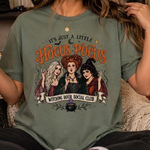 May include: An olive green t-shirt featuring a graphic print of three witches with the text "IT'S JUST A LITTLE HOCUS POCUS" and "WITCHING HOUR SOCIAL CLUB" above and below the illustration, which also includes a small black cauldron with purple smoke.