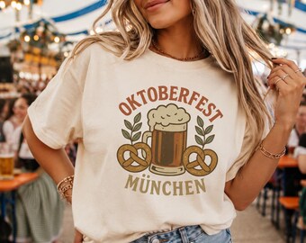 Puede incluir: Una camiseta de color crema con un estampado gráfico de una jarra de cerveza con espuma, dos pretzels y ramas verdes. El texto dice OKTOBERFEST en una fuente marrón curva sobre la jarra y MÜNCHEN en una fuente marrón recta bajo el gráfico.