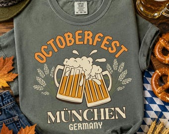 Puede incluir: Una camiseta gris carbón con un gráfico de dos jarras de cerveza chocando con espuma derramándose. El texto dice OCTOBERFEST sobre las jarras y MÜNCHEN GERMANY debajo. El diseño incluye espigas de trigo a ambos lados de las jarras.