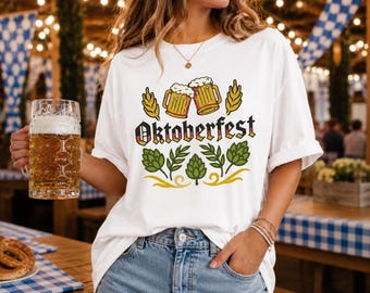 Puede incluir: Una camiseta blanca con un estampado gráfico que muestra el texto Oktoberfest en letras negras con un patrón de bandera alemana. El diseño incluye dos jarras de cerveza, espigas de trigo amarillas y conos de lúpulo verdes.