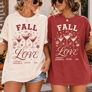 May include: A cream-colored t-shirt and a brick-red t-shirt, both featuring the text "FALL in Love" in a decorative font. The graphic includes three cocktail glasses surrounded by autumn leaves and stars, with "Stephanie's Bachelorette", "Location", and "2026" printed below.