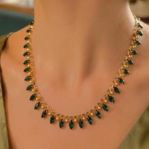 以下が含まれることがあります： エメラルドグリーンの涙型の石が付いたゴールドトーンのネックレス。グリーンのサテンのトップスに合わせて着用されています。この繊細なジュエリーは、特別な機会に最適です。