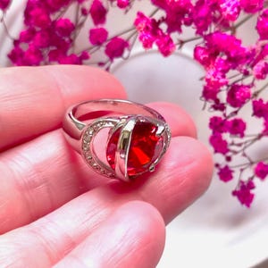 May include: A silver ring with a large, square, red gemstone as the focal point. The ring has a double band design with a halo of small, clear stones around the gemstone. The ring is displayed against a backdrop of pink flowers.