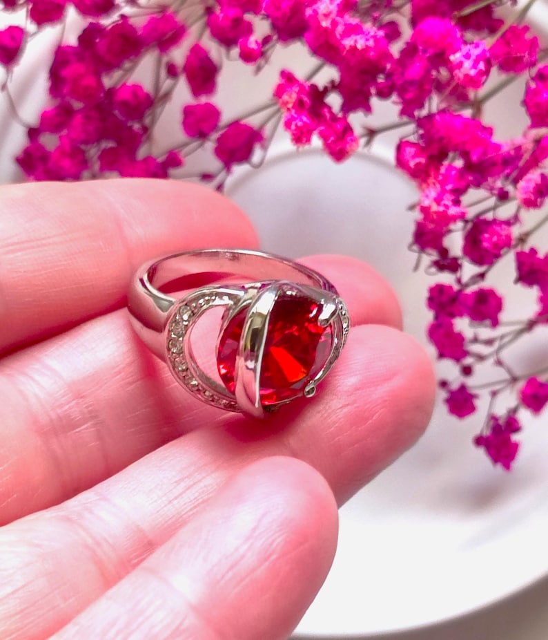 May include: A silver ring with a large, square, red gemstone as the focal point. The ring has a double band design with a halo of small, clear stones around the gemstone. The ring is displayed against a backdrop of pink flowers.