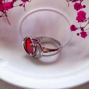May include: A silver ring with a large, oval-shaped, red gemstone as the centerpiece. The gemstone is surrounded by a silver setting with a small, decorative design. The ring is set on a white surface, with pink flowers in the background.