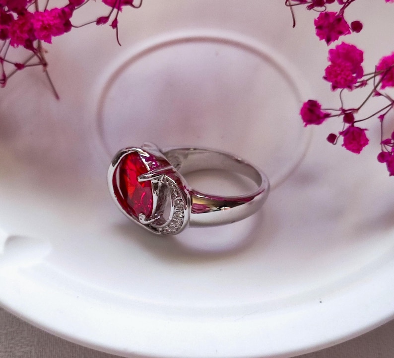 May include: A silver ring with a large, oval-shaped, red gemstone as the centerpiece. The gemstone is surrounded by a silver setting with a small, decorative design. The ring is set on a white surface, with pink flowers in the background.
