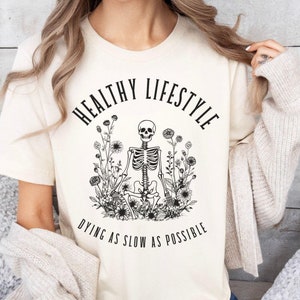 May include: A beige t-shirt with a black graphic of a skeleton meditating in a circle of flowers. The text "Healthy Lifestyle Dying as Slow as Possible" is printed below the graphic.