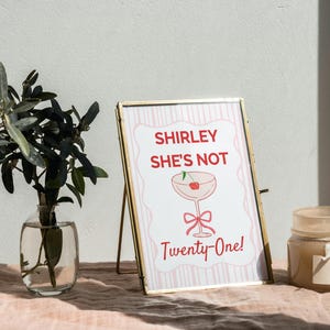 May include: A framed print with the text "Shirley She's Not Twenty-One!" in red and white, featuring a cocktail glass illustration. The frame is gold, and the print is displayed on a pink surface with a vase of greenery and a candle.