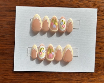May include: A set of press-on nails featuring a light peach base with a white lace design at the cuticle. Some nails have a floral design with pink, yellow, and blue flowers. The nails are oval-shaped and arranged on a white card.