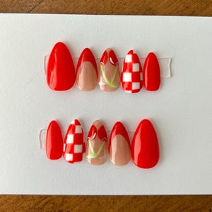 May include: Set of ten red and white press-on nails with different designs. Some nails have a red base with a white checkered pattern, others have a white base with a red cherry design, and some are solid red.
