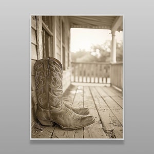 May include: A pair of brown cowboy boots with intricate stitching on the leather are sitting on a wooden porch. The boots are worn and have a rustic look. The porch is made of wood planks and has a railing in the background.
