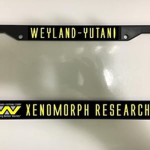 Könnte beinhalten: Schwarzer Kennzeichenrahmen mit gelbem Text "WEYLAND-YUTANI" oben und "XENOMORPH RESEARCH" unten. Das Weyland-Yutani Corp Logo ist ebenfalls sichtbar. Der Rahmen ist rechteckig und hat einen schwarzen Rand.