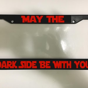 May include: Black license plate frame with the red text "MAY THE DARK SIDE BE WITH YOU". The frame is rectangular and has a clear center for the license plate.