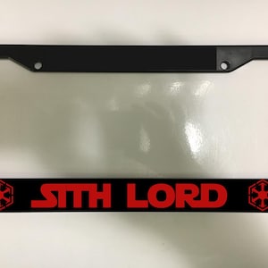 Puede incluir: Marco negro para matrícula con las palabras "SITH LORD" en rojo. El marco tiene un borde negro y una inserción de plástico transparente. El símbolo Sith está a cada lado del texto.