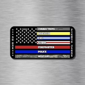 May include: Rectangular black license plate featuring a stylized American flag design. The flag incorporates stripes representing various professions: Corrections, Dispatch, EMS, Nurse, Firefighter, Police, and Military. The text "NONE FIGHTS ALONE" is displayed.