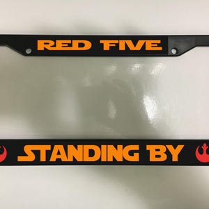 May include: Black license plate frame with orange text that reads "RED FIVE" at the top and "STANDING BY" at the bottom. The frame has a clear plastic insert and a red Rebel Alliance symbol on each side of the bottom text.