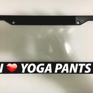 May include: Black license plate frame with the text "I ❤️ YOGA PANTS" in white letters and a red heart. The frame is rectangular with rounded corners and has mounting holes.